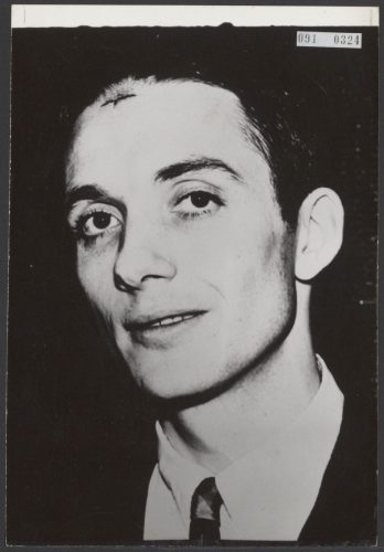 Portret van een jonge man met donker, achterovergekamd haar, wit overhemd en stropdas. Op zijn voorhoofd, vlak onder de haargrens, een klein donker litteken.