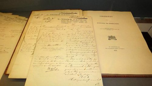 Handgeschreven vellen met de gedrukte aanhef Wij Willem II, bij de gratie Gods, Koning der Nederlanden liggen in een vitrine naast een opengeslagen gedrukte titelpagina Grondwet voor het Koningrijk der Nederlanden, 1848.